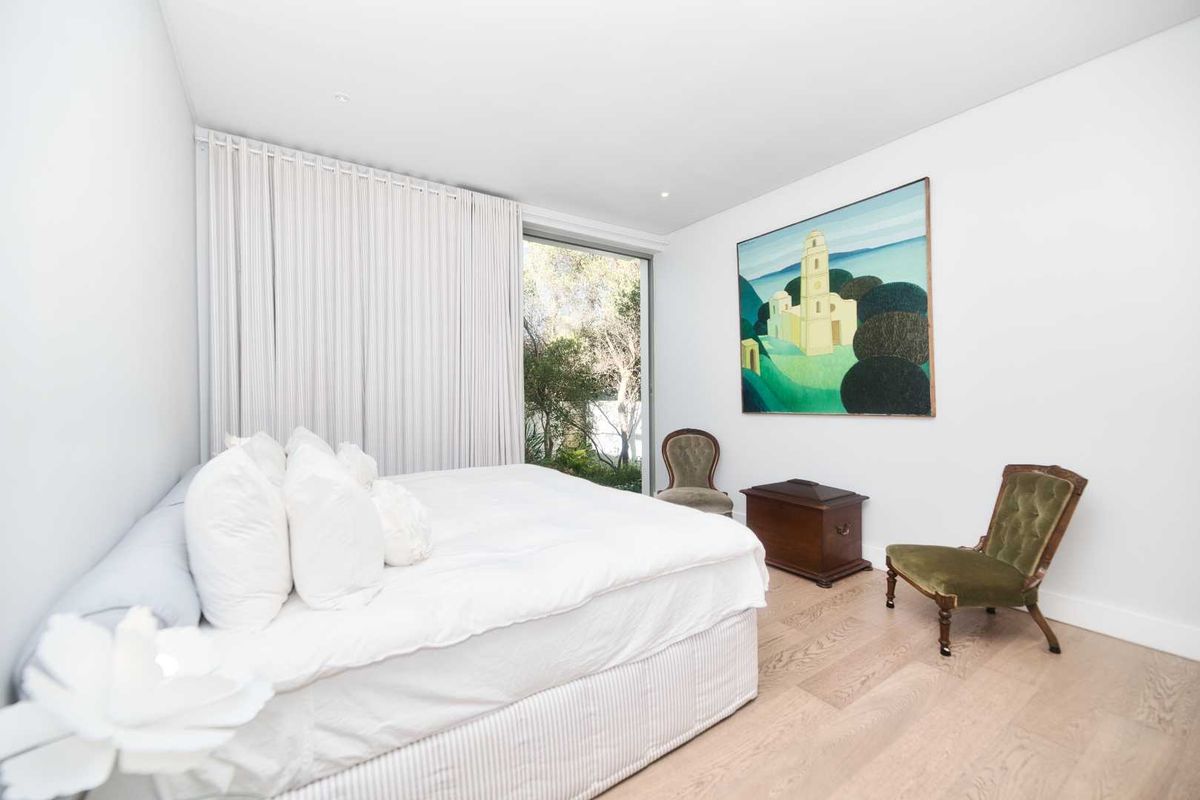 A bright and modern bedroom featuring light timber flooring, recessed ceiling lighting, and floor-to-ceiling glass sliding doors opening to a private garden view.