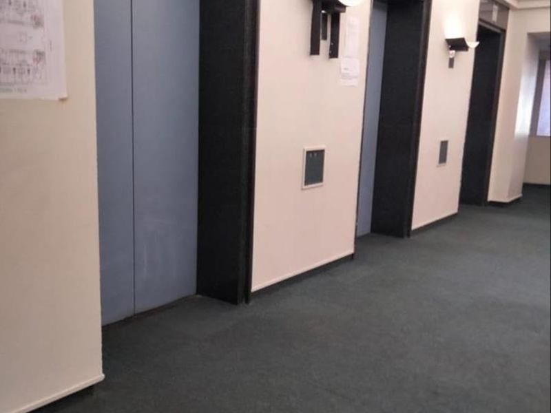 A communal apartment building hallway featuring multiple elevator access points, wall-mounted lighting, and carpeted flooring.