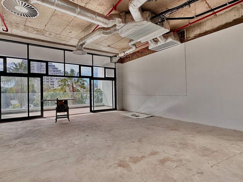 Spacious open-plan office area featuring industrial-style exposed concrete ceilings, ductwork, and large floor-to-ceiling windows leading to a private balcony.