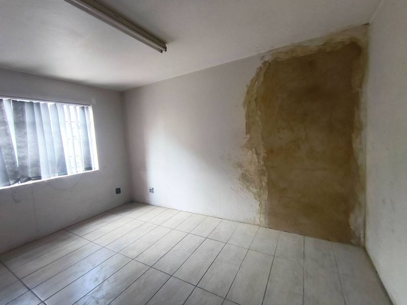 An empty office space featuring tiled flooring, vertical blinds, and a fluorescent light fixture, currently requiring wall surface repairs and finishing.