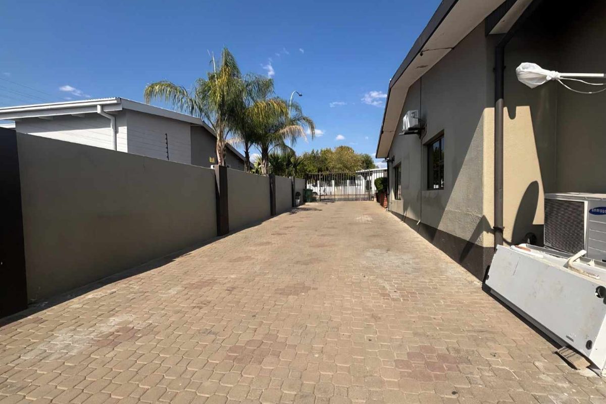 A paved side driveway featuring a high boundary wall, security gate access, and external building infrastructure including an air conditioning unit.
