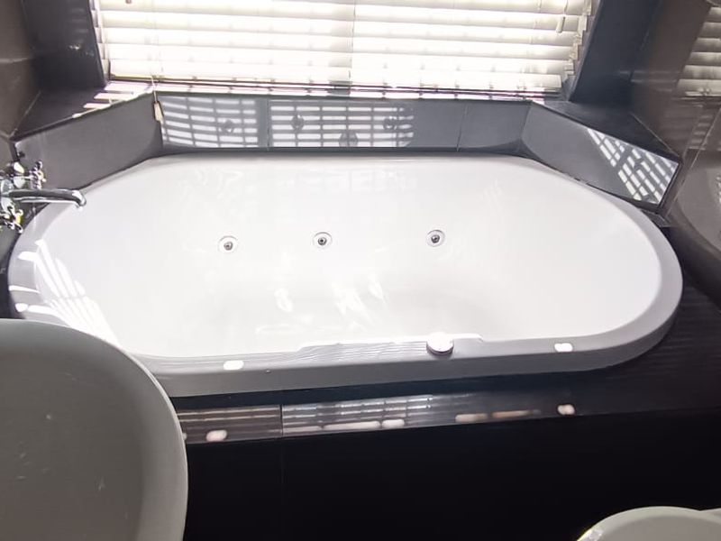 A modern bathroom featuring a built-in jetted bathtub, a basin, and a toilet, finished with dark tiled walls and venetian blinds.