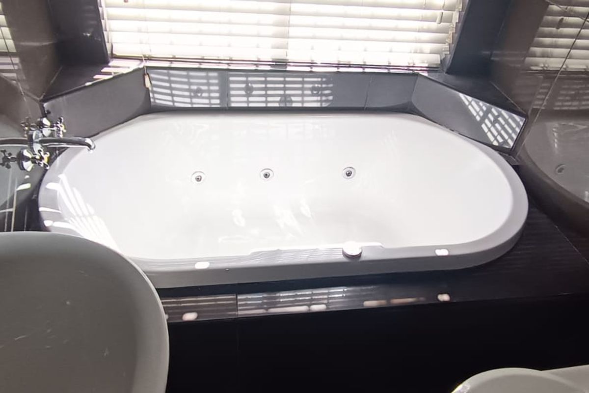 A modern bathroom featuring a built-in jetted bathtub, a basin, and a toilet, finished with dark tiled walls and venetian blinds.