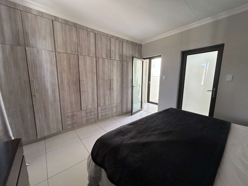 A well-maintained bedroom featuring extensive built-in wooden cupboards, neutral tiled flooring, and direct access to an outdoor area through a glass door.