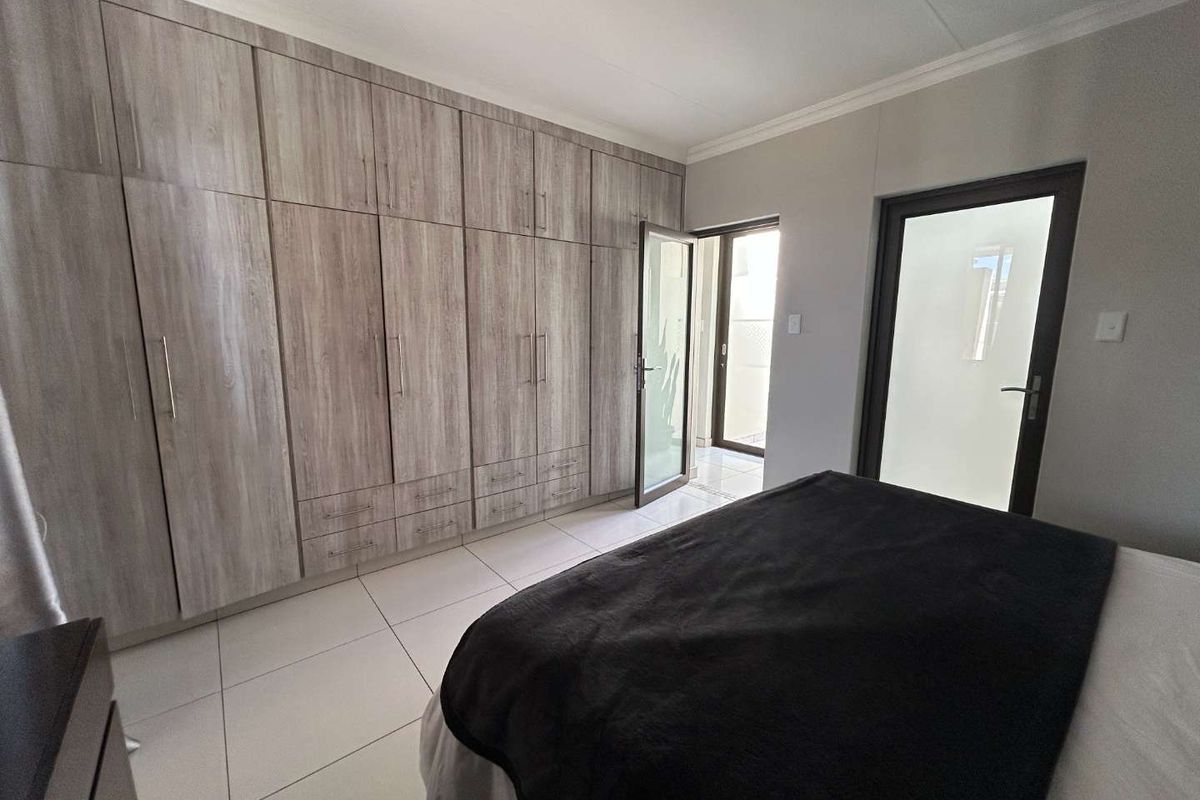 A well-maintained bedroom featuring extensive built-in wooden cupboards, neutral tiled flooring, and direct access to an outdoor area through a glass door.