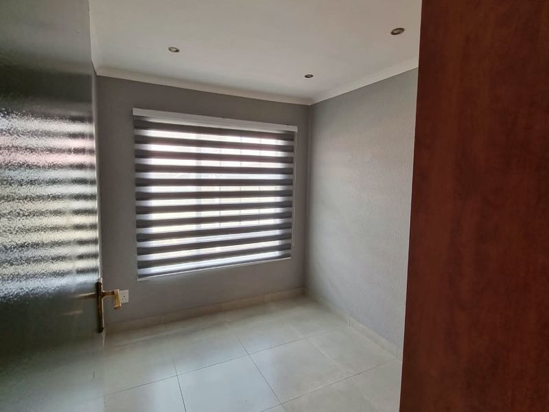 A clean, empty bedroom featuring neutral grey walls, tiled flooring, recessed downlights, and modern roller blinds.