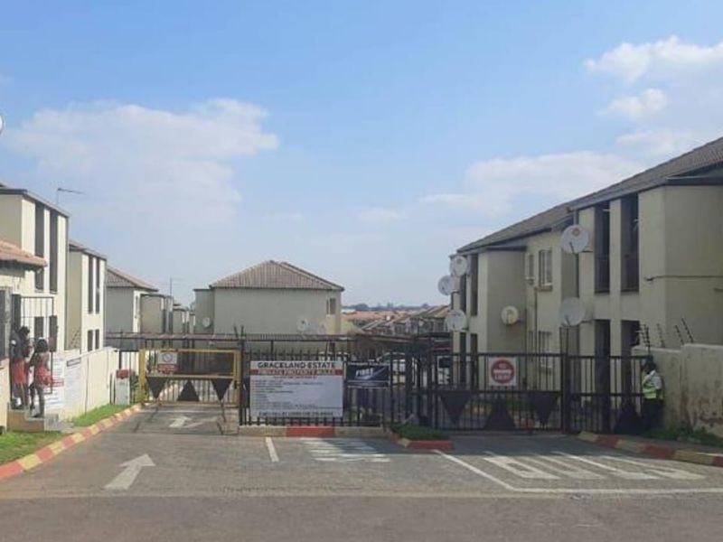 Street view of the residential estate entrance featuring a security gate, paved driveway, and multi-unit townhouse buildings with tiled roofs.