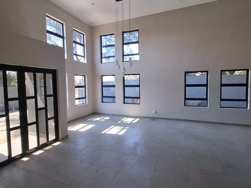 Spacious double-volume living area featuring large format floor tiles, abundant natural light through multiple aluminium-framed windows, and a modern glass-panelled door.