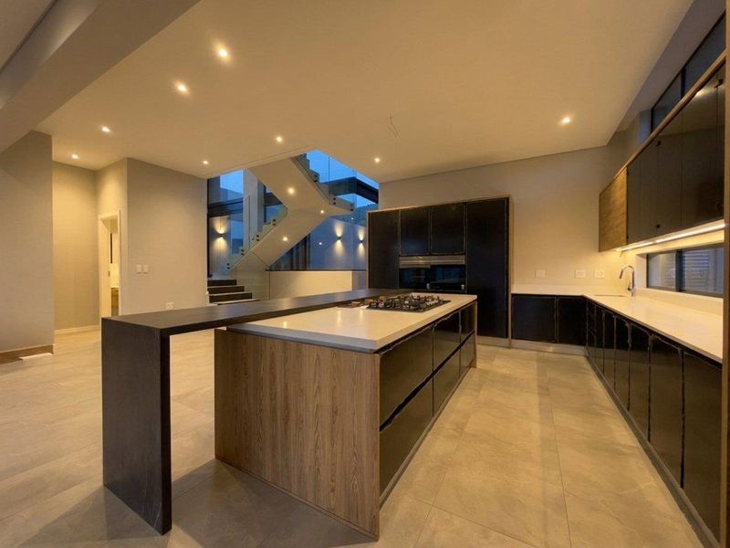 Modern open-plan kitchen featuring a large island with gas hob, sleek cabinetry, stone countertops, and integrated appliances, adjacent to a contemporary staircase.