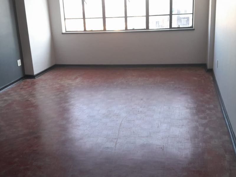 An empty living room featuring original parquet flooring, large windows providing natural light, and neutral wall finishes.