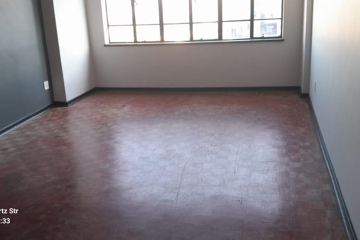 An empty living room featuring original parquet flooring, large windows providing natural light, and neutral wall finishes.