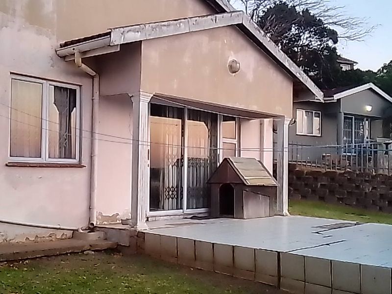 Rear exterior view of the townhouse featuring a tiled patio, sliding glass doors, and a retaining wall, showing signs of weathered exterior paint.