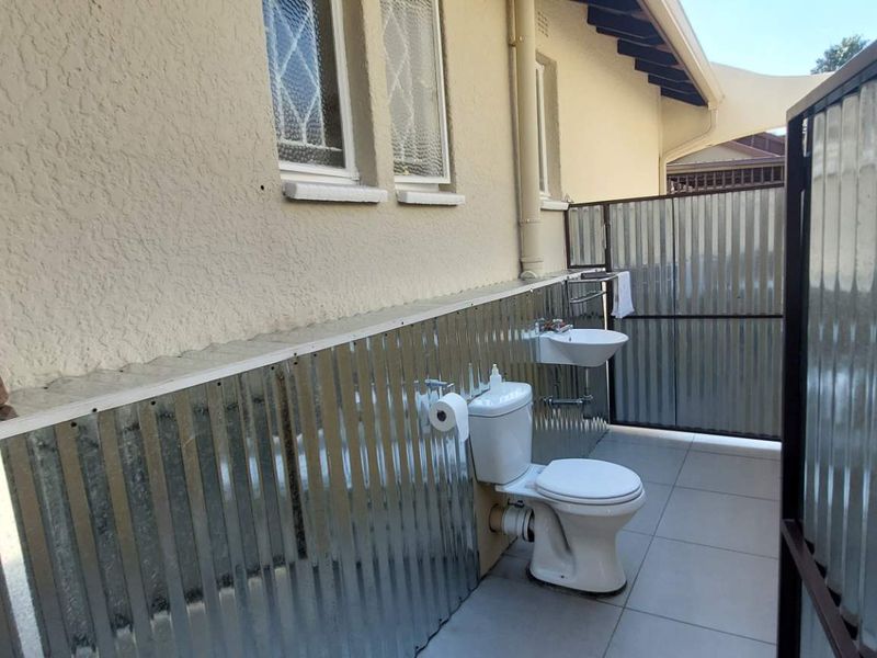 An outdoor bathroom area featuring a toilet, wall-mounted basin, tiled flooring, and corrugated metal privacy screening.