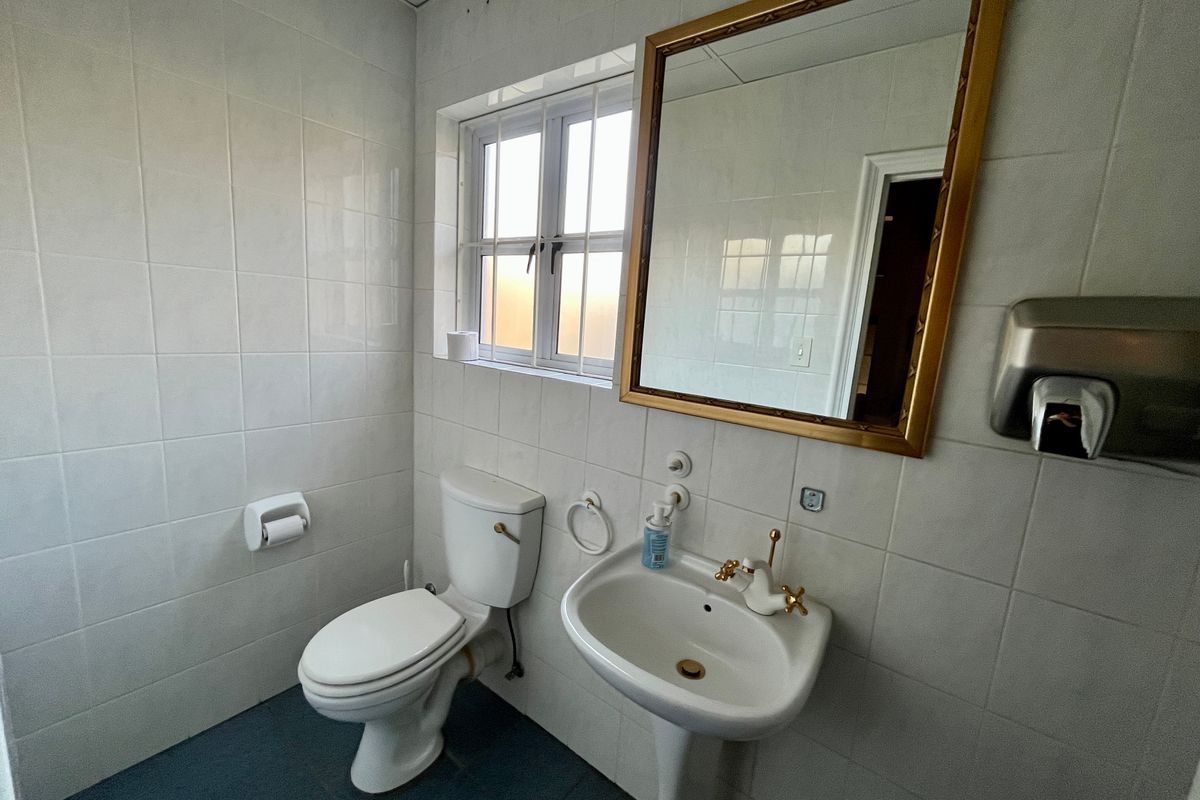 A functional bathroom featuring a pedestal basin, toilet, and wall-mounted soap dispenser, finished with white wall tiles and a window with security bars.