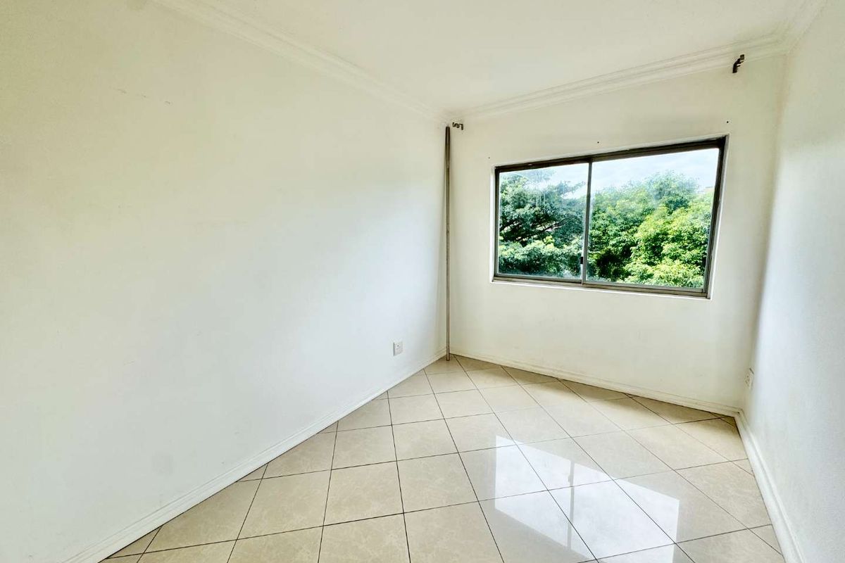 A bright, empty bedroom featuring neutral tiled flooring, white walls, and a large window with a pleasant green outlook.