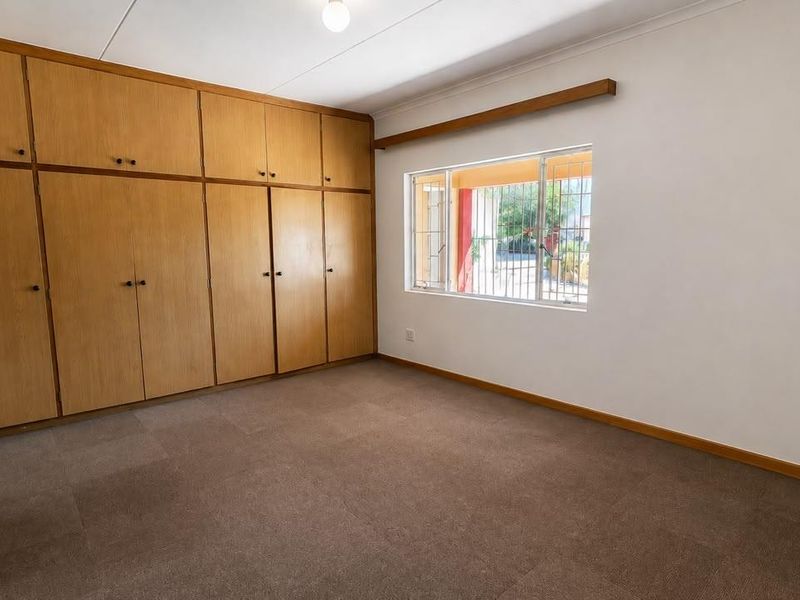 A spacious, empty bedroom featuring extensive built-in wooden cupboards, carpeted flooring, and a window fitted with security bars.
