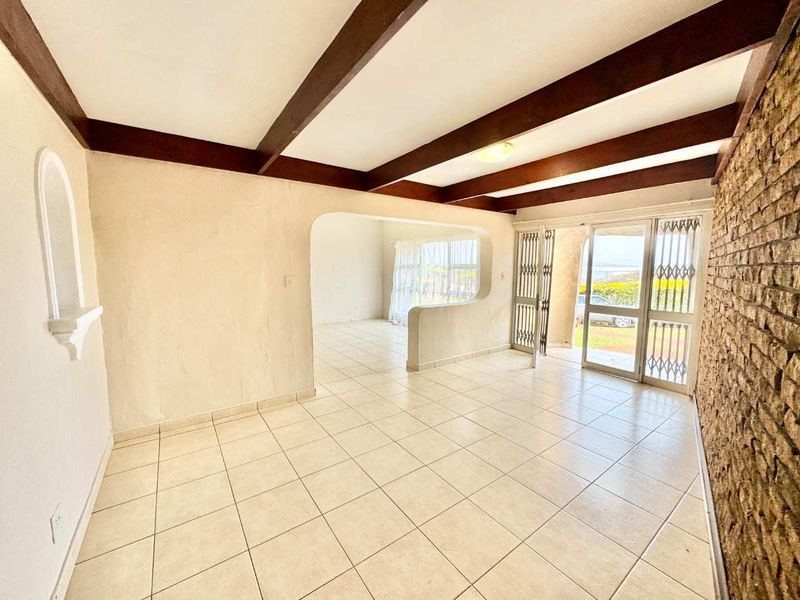 Spacious living area featuring exposed timber ceiling beams, a rustic brick feature wall, and neutral ceramic floor tiles with access to an outdoor area.