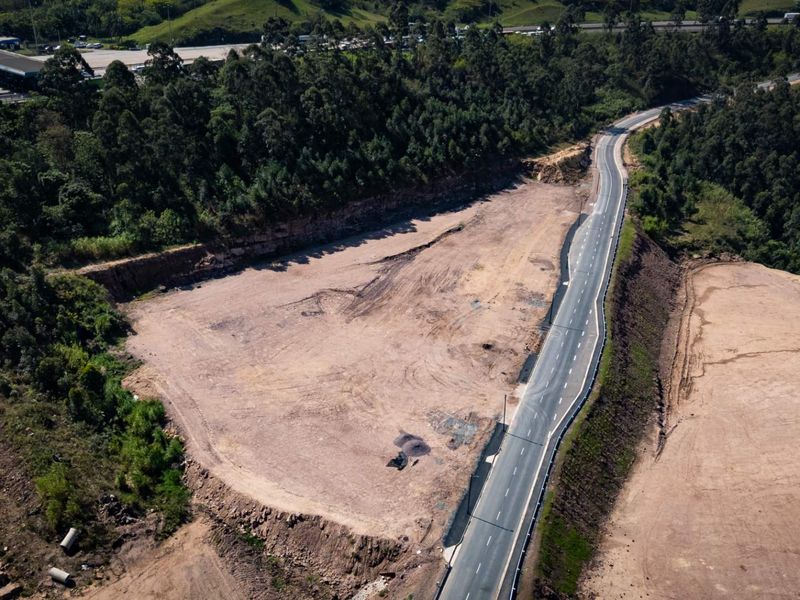 Aerial view of a cleared industrial land plot adjacent to a paved road with guardrails, surrounded by dense forest.
