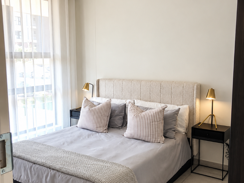 A well-lit bedroom featuring neutral walls, tiled flooring, and a large window with sheer curtains, creating a bright and comfortable living space.