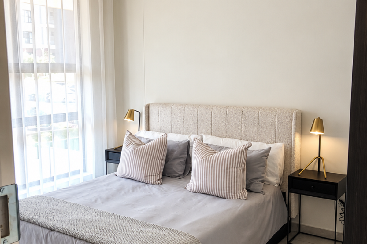 A well-lit bedroom featuring neutral walls, tiled flooring, and a large window with sheer curtains, creating a bright and comfortable living space.
