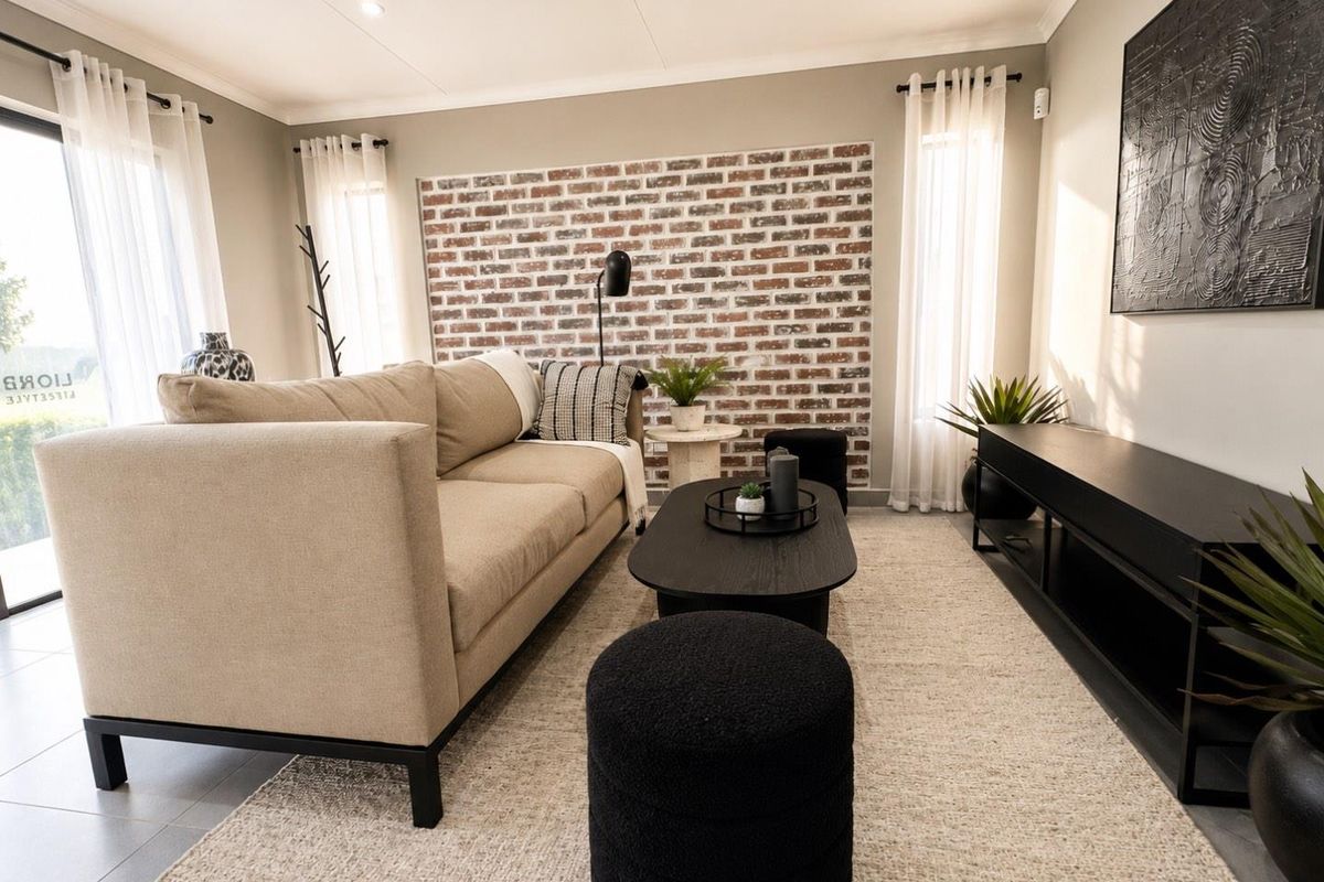 A well-lit living room featuring a decorative exposed brick feature wall, tiled flooring, and large windows with sheer curtains.