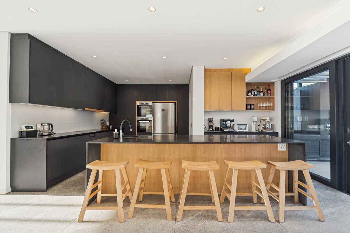 Modern kitchen featuring sleek dark cabinetry, a large wooden island with stone countertop, integrated appliances, and recessed lighting, opening to an outdoor area.