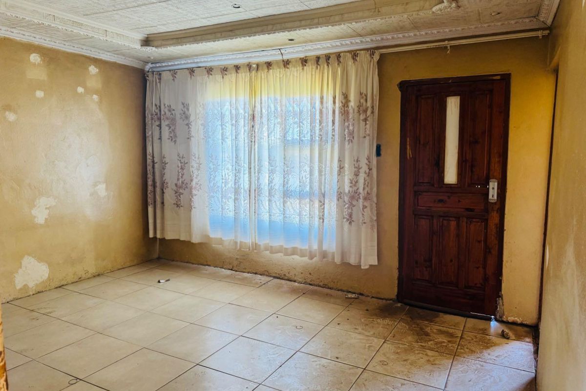 An empty living room featuring tiled flooring, a wooden door, and a decorative ceiling, requiring cosmetic updates and wall repairs.