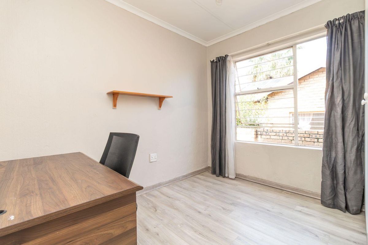 A functional bedroom featuring light-toned laminate flooring, a window with security bars, and a wall-mounted shelf.