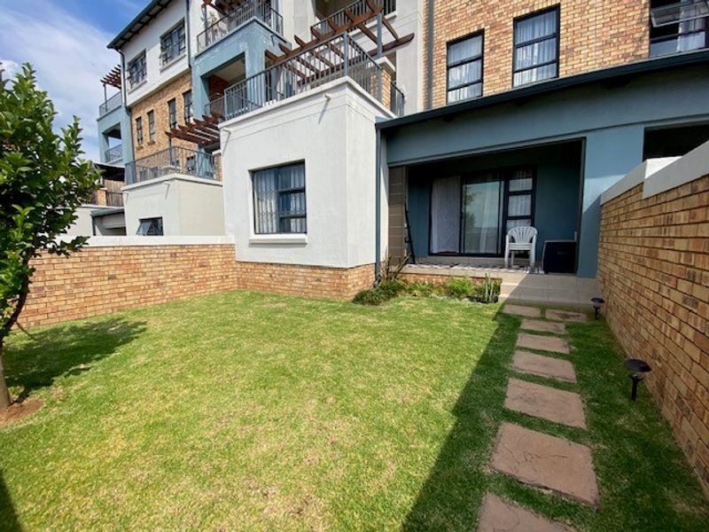 A private rear garden featuring a well-maintained lawn, paved pathway, and a covered patio area attached to the apartment building.