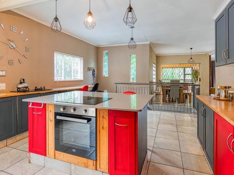 Open-plan kitchen featuring a central island with built-in oven and cooktop, contrasting cabinetry, and tiled flooring, overlooking a dining area with metal balustrade.