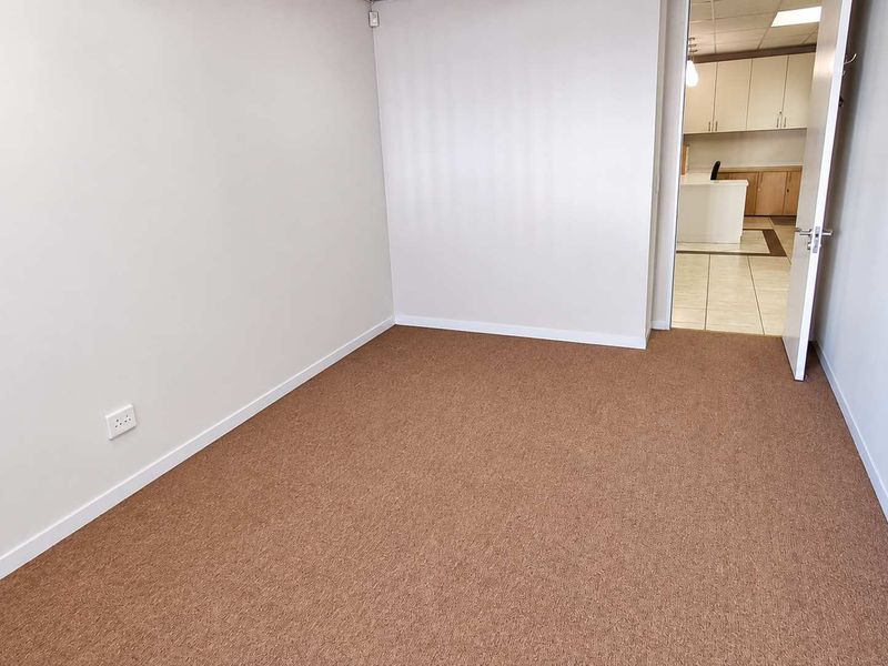 A clean, empty office space featuring neutral walls and carpet flooring, with a doorway leading to an adjacent kitchenette area.