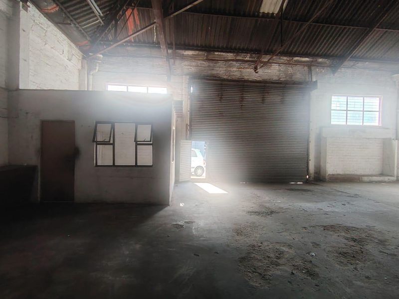 Spacious industrial warehouse unit featuring high ceilings with exposed steel trusses, concrete flooring, a roller shutter door, and an integrated internal office partition.