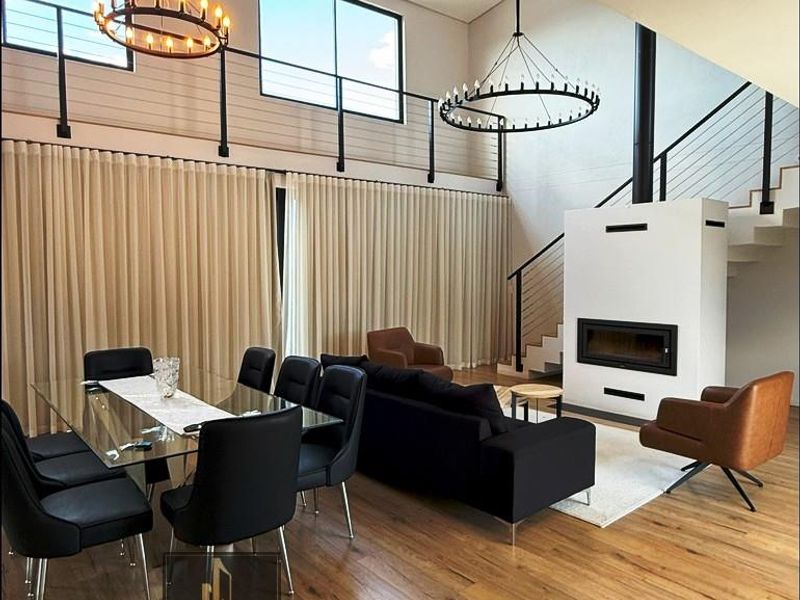 Spacious open-plan living and dining area featuring double volume ceilings, a modern built-in fireplace, timber flooring, and a mezzanine level with metal railings.