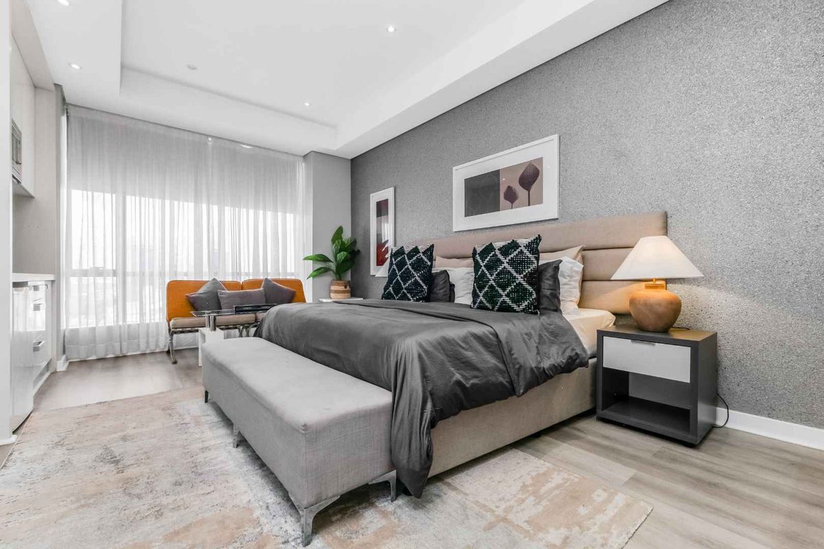 A modern, well-lit bedroom featuring a textured feature wall, recessed ceiling lighting, and large windows with sheer curtains.