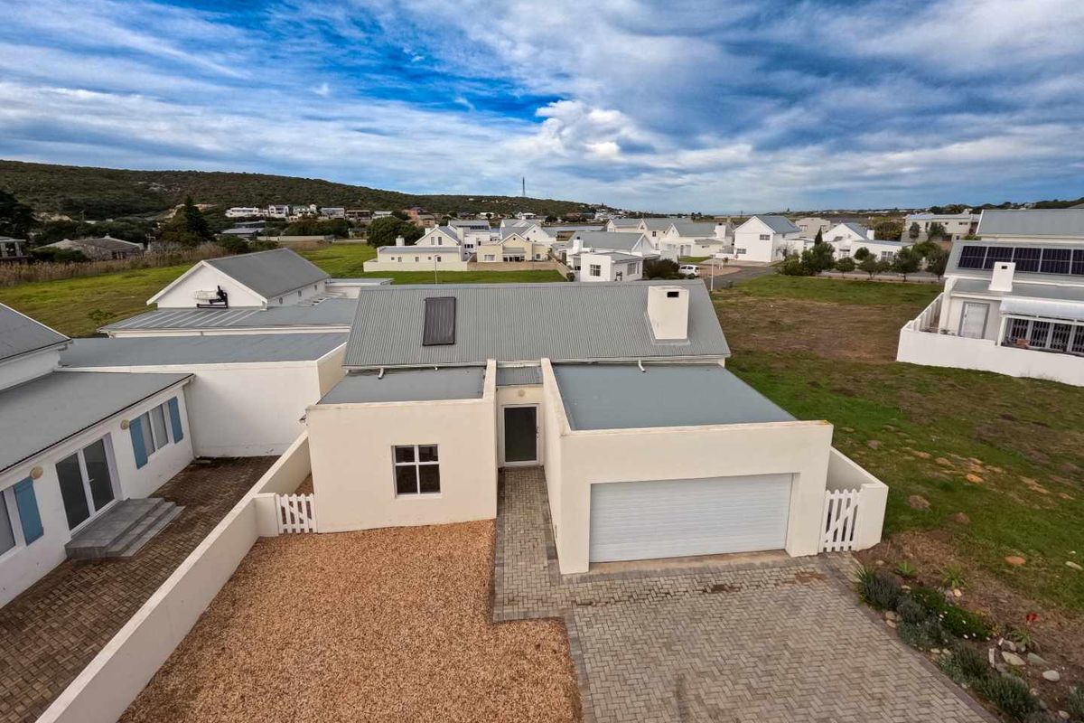 Aerial view of a modern residential property featuring a corrugated metal roof, paved driveway, attached garage, and gravel front yard in a coastal estate.