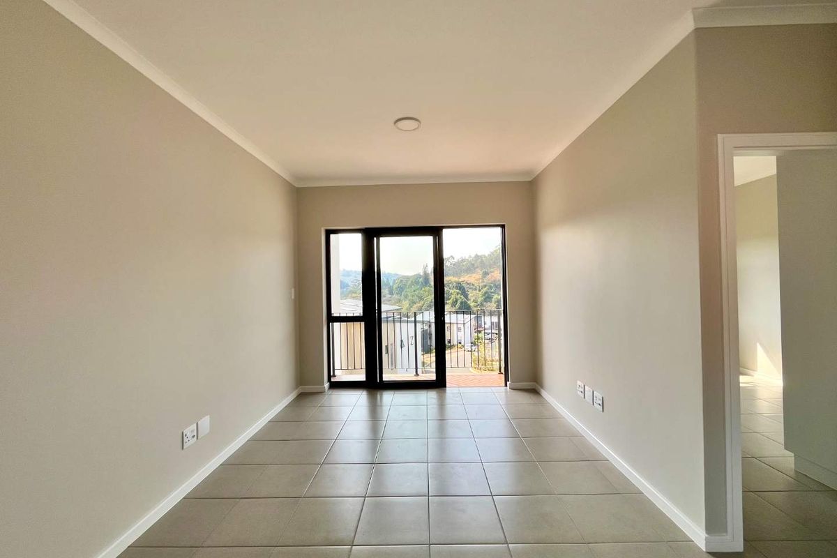 A bright, empty living area featuring neutral tiled floors, clean walls, and a sliding glass door providing access to a balcony with scenic views.