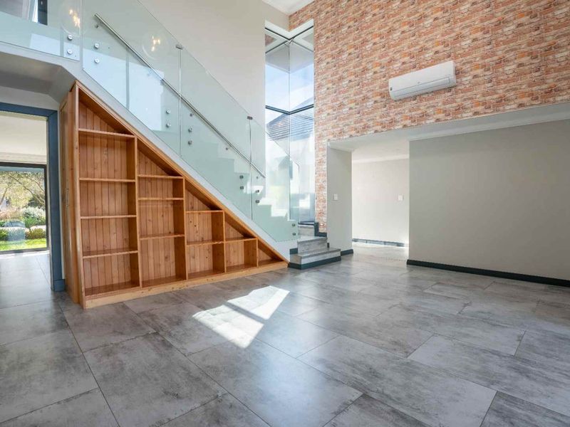 Spacious living area featuring a modern glass staircase, built-in under-stair shelving, and an exposed brick feature wall with tiled flooring throughout.