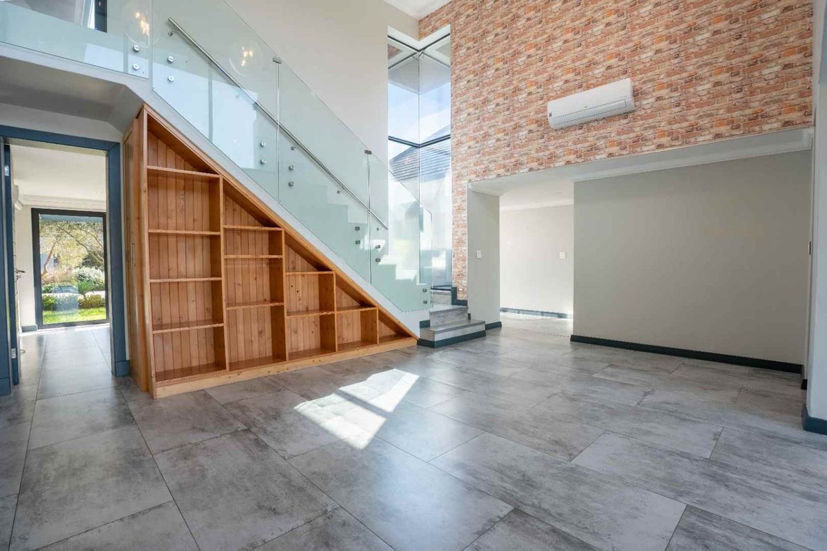Spacious living area featuring a modern glass staircase, built-in under-stair shelving, and an exposed brick feature wall with tiled flooring throughout.