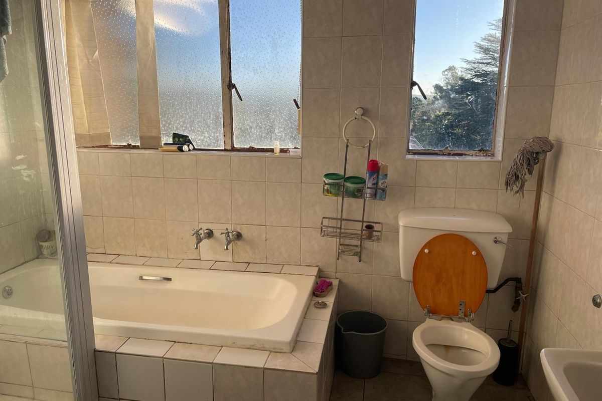 A dated bathroom featuring a built-in bathtub, toilet, and tiled walls with large windows providing natural light.
