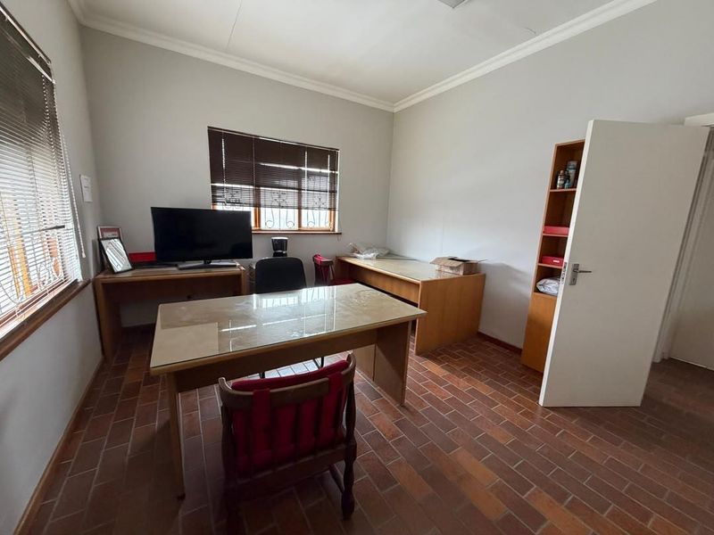 A functional office space featuring terracotta tile flooring, neutral walls, and natural light provided by two windows with fitted venetian blinds.