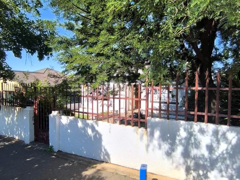 Street view of the property frontage featuring a masonry boundary wall with metal fencing and mature trees providing partial shade.