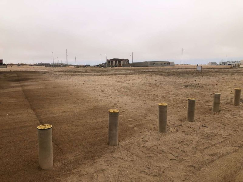 A view of a vacant residential plot featuring concrete bollards and surrounding neighborhood infrastructure under an overcast sky.