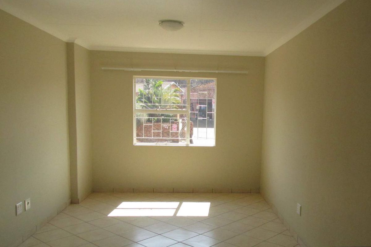 A bright, empty living area featuring neutral tiled flooring, a central ceiling light, and a window with security bars.