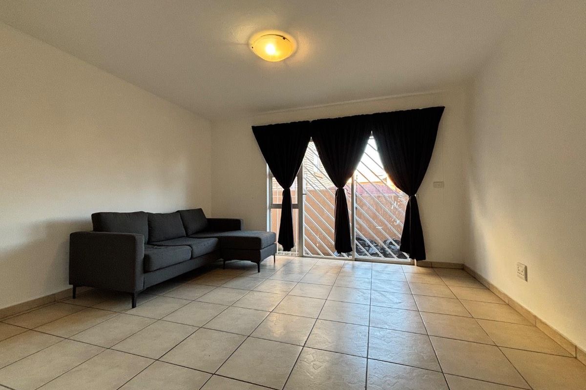 A simple living room featuring neutral floor tiles, a flush-mount ceiling light, and a sliding glass door with security bars leading to the exterior.