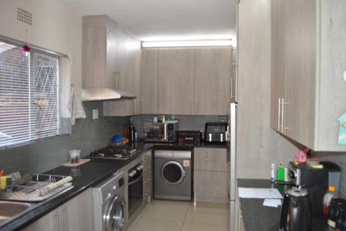 A functional galley-style kitchen featuring light wood-grain cabinetry, dark stone countertops, a built-in gas hob, extractor fan, and integrated laundry appliance space.