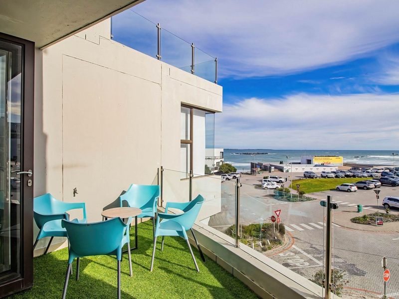 A modern balcony featuring a glass balustrade, artificial turf flooring, and an outdoor tap, offering scenic coastal and street views.