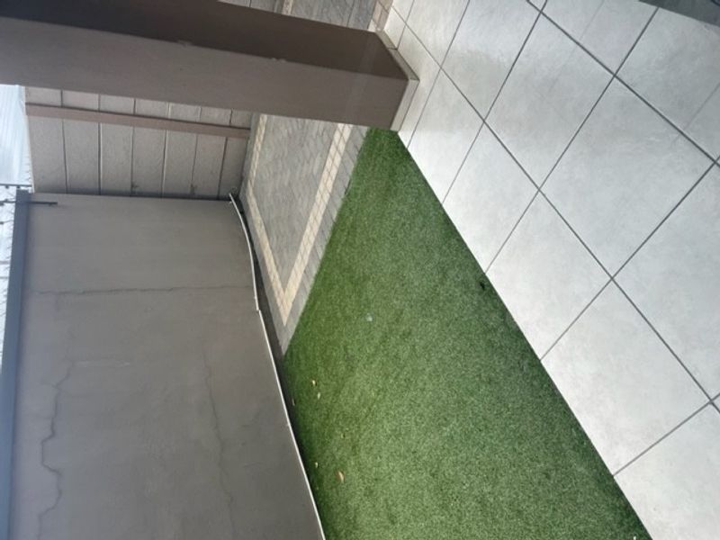 A compact outdoor patio area featuring tiled flooring, a section of artificial grass, and paved detailing, suitable for a small seating arrangement.
