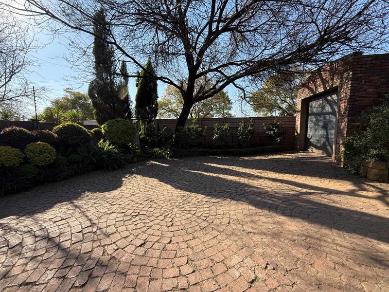A well-maintained front exterior featuring a circular pattern cobblestone driveway leading to a brick garage, surrounded by mature trees and established landscaping.