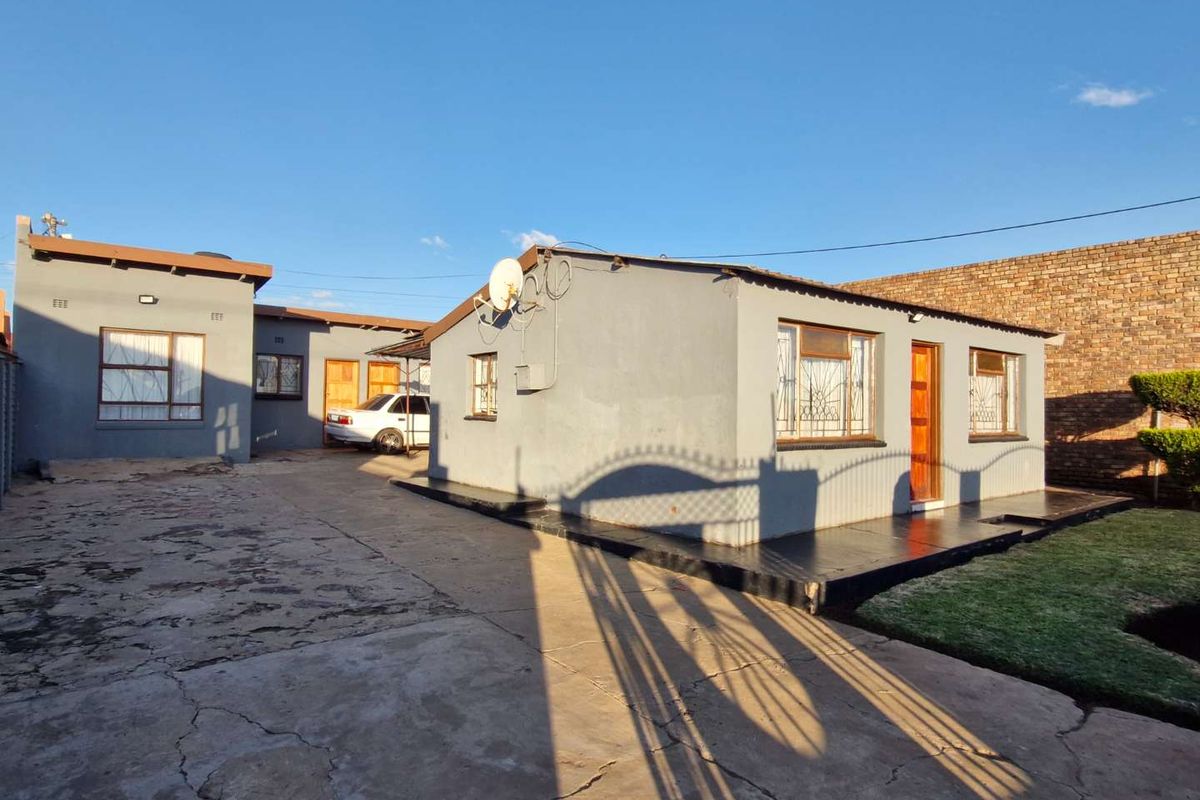Exterior view of a residential property featuring a concrete driveway, brick boundary wall, and multiple building structures with metal-framed windows.