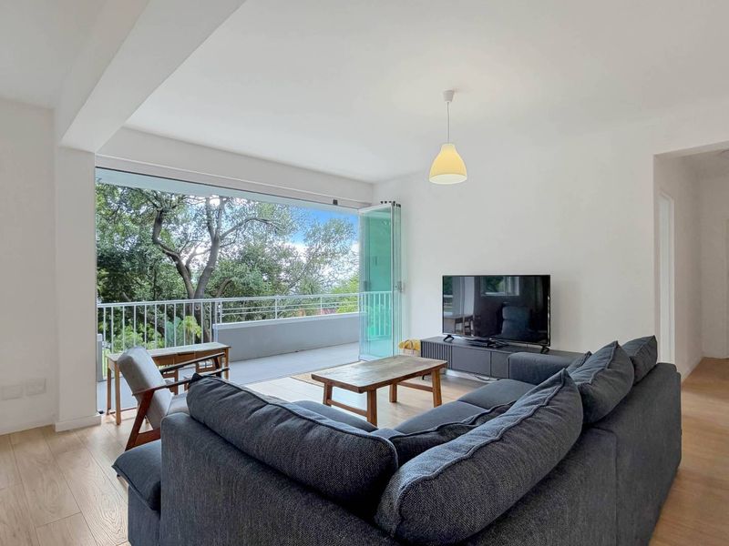 Bright living room featuring light wood-look flooring and large bi-fold doors opening onto a private balcony with leafy views.